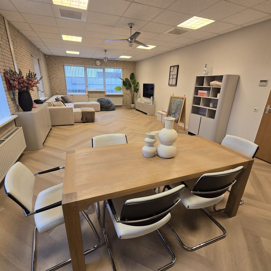 Ruime kamer met een houten vergadertafel en stoelen, en op de achtergrond een zithoek met een bank, een zitzak en een televisie.