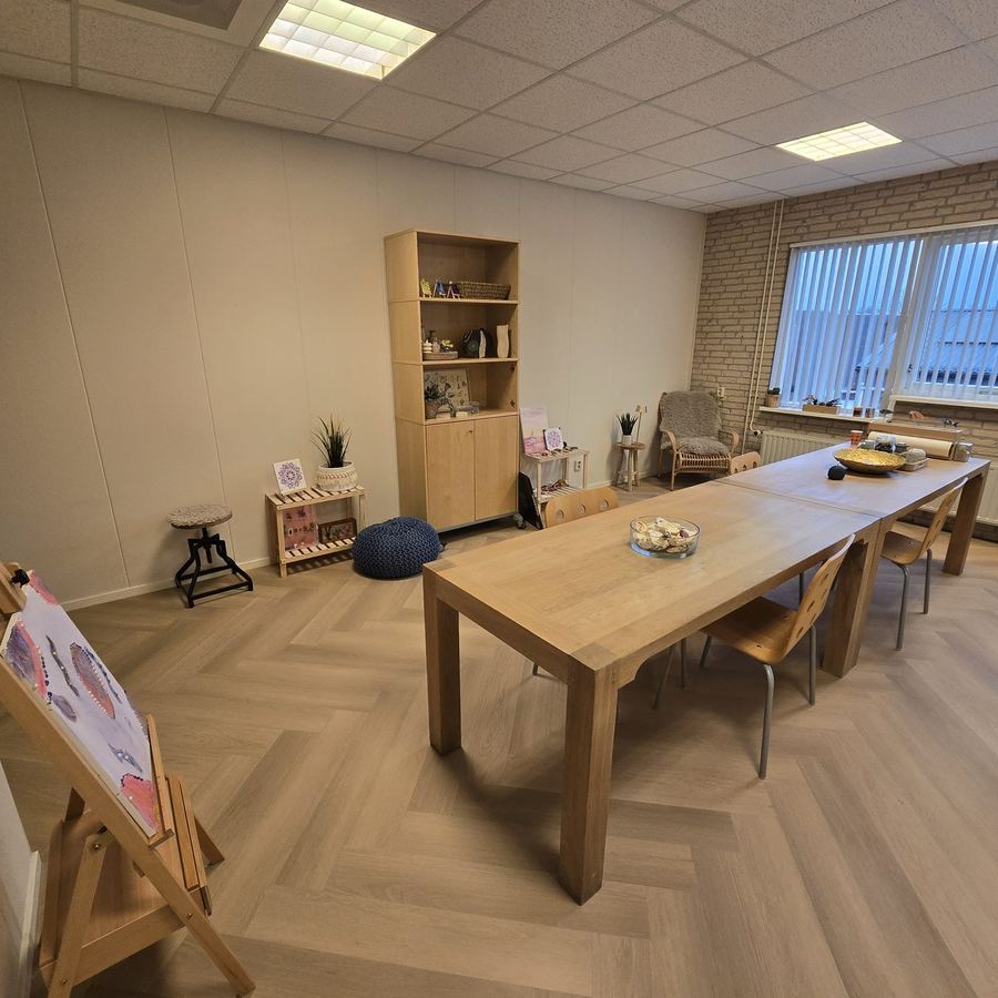 Ruimte met een lange tafel, een schildersezel en open kasten met materialen.