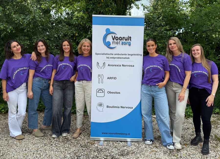 Zeven medewerkers in paarse shirts van World Eating Disorders Action Day staan naast een banner van Vooruit met Zorg.