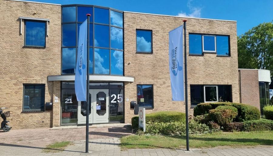 Bakstenen kantoorpand met twee vlaggen van Vooruit met Zorg bij de ingang; naast de deur staan de huisnummers 21 en 25.