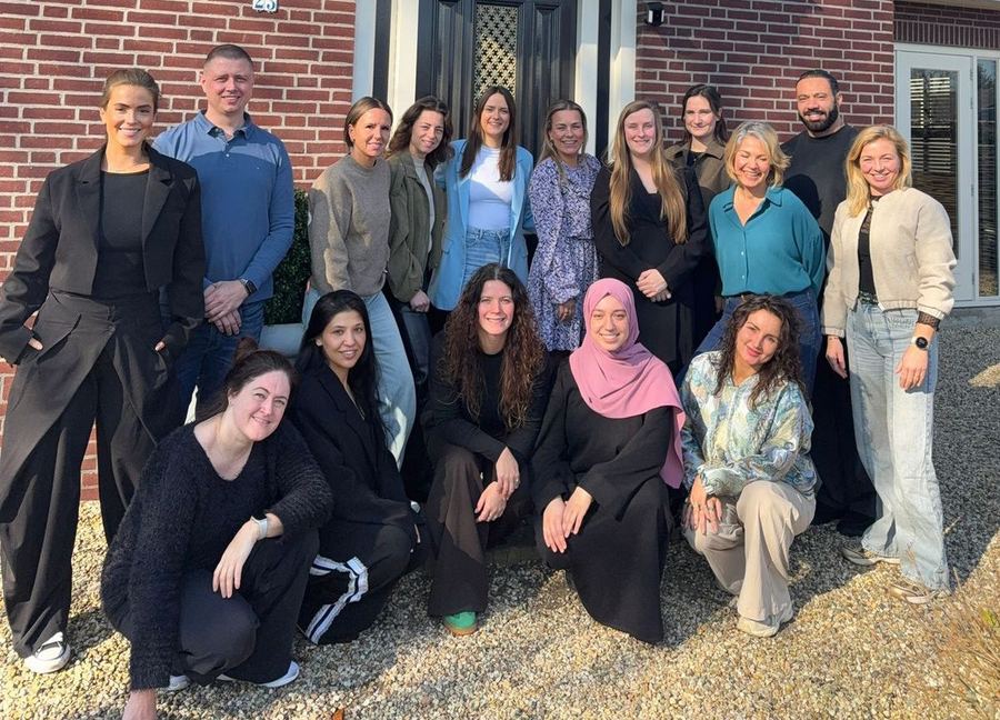 Groepsfoto van het team van Vooruit met Zorg voor een bakstenen gebouw: veertien mensen staan, vijf zitten gehurkt op het grind ervoor.