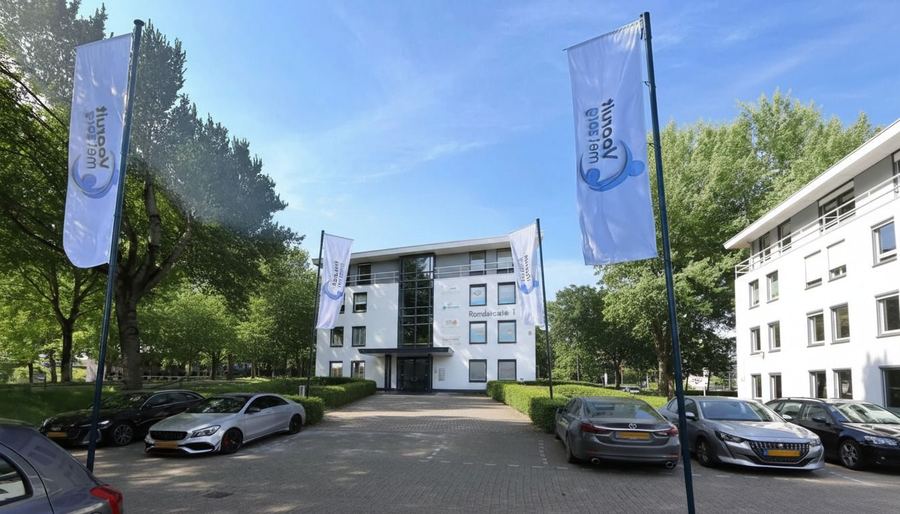 Wit kantoorgebouw met vier vlaggen van Vooruit met Zorg langs de oprit en parkeerplaatsen ervoor.